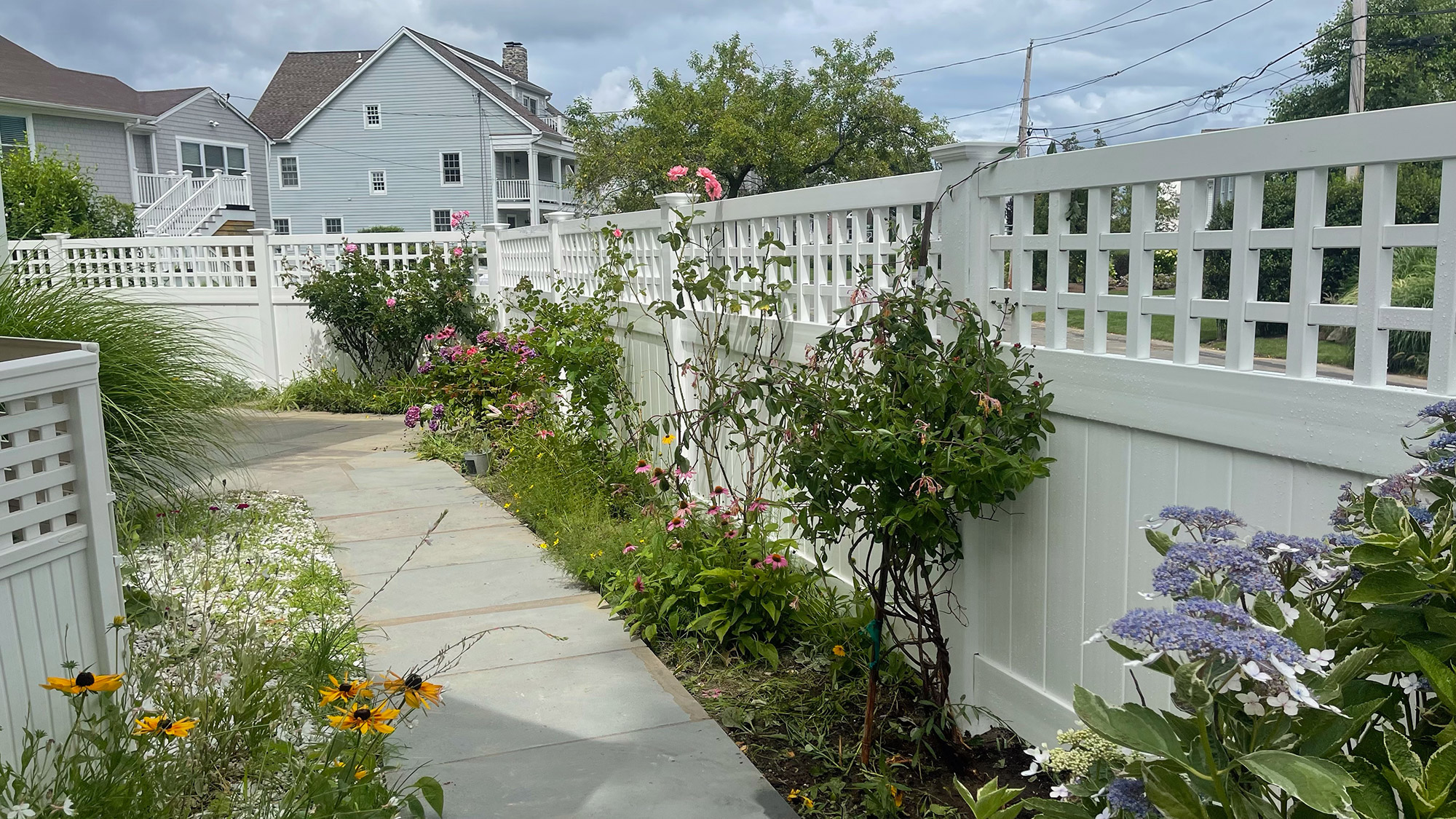 A walkway with flowers and a white fence.