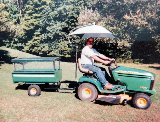 man riding a lawn mower with a trailer and an umbrella.